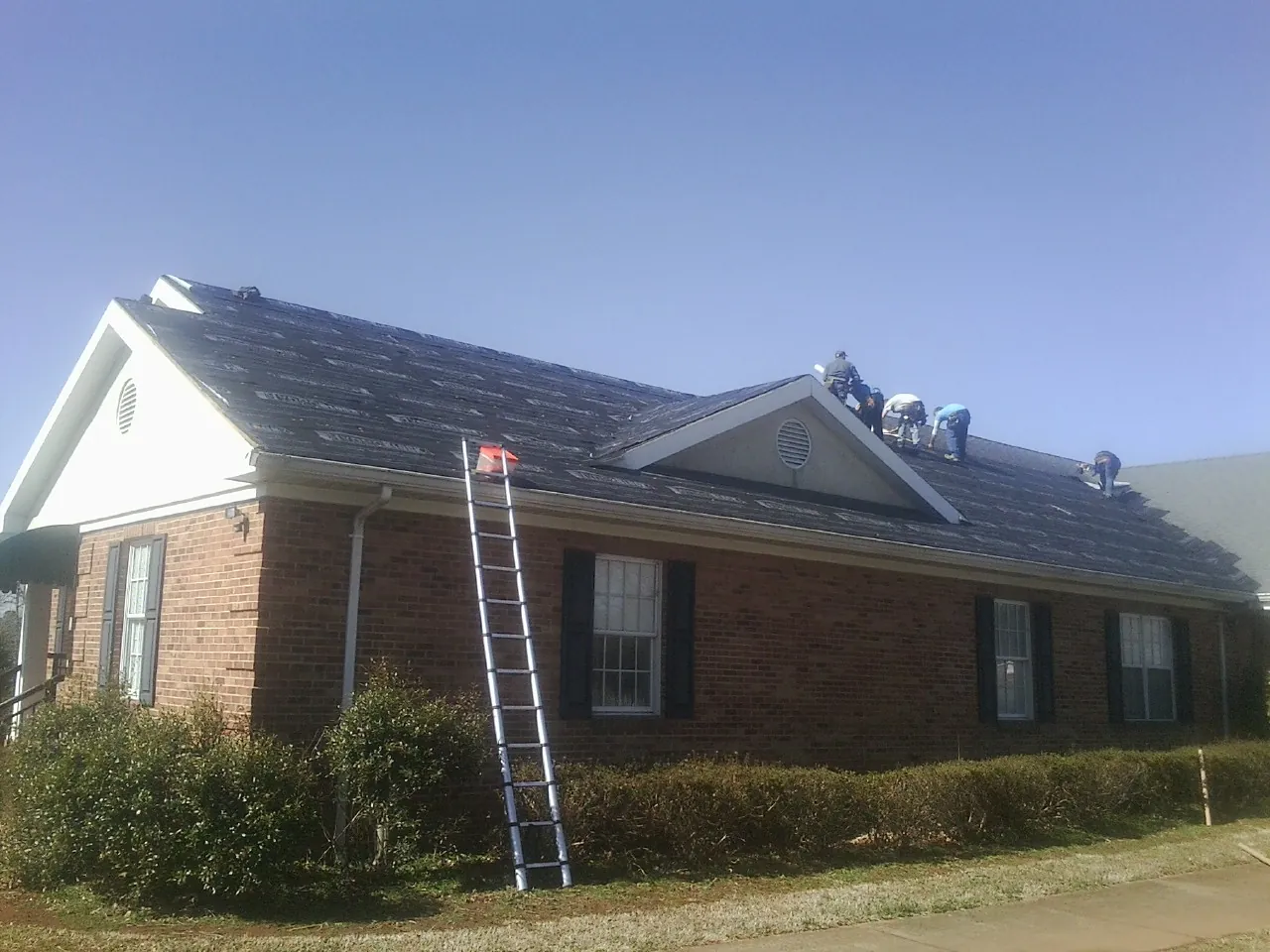 Crew tearing shingles off a brick ranch roof with a ladder set — Emergency Roof Repair work by our crew in Tropic
