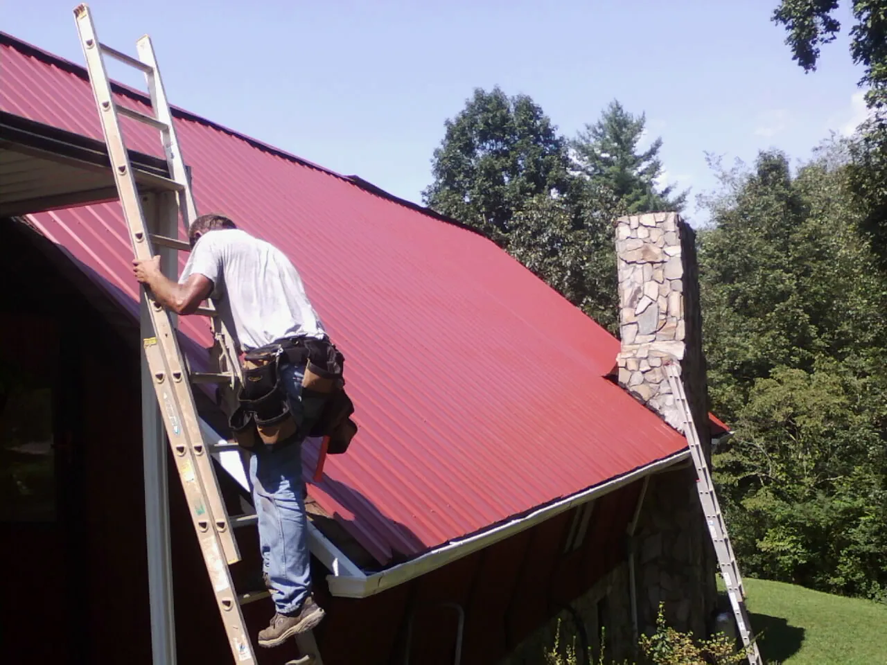 Roofer on a ladder beside a red metal roof and stone chimney — Soffit & Fascia Repair by Roofing Brothers in Tropic